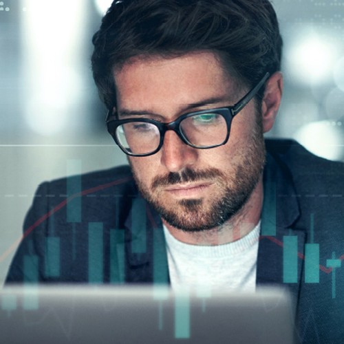A man in glasses focuses intently on a laptop, analyzing data overlaid with digital graphs and charts in a modern, softly lit office environment.
