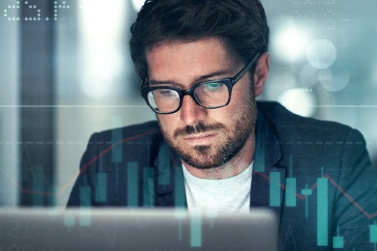 A man in glasses focuses intently on a laptop, analyzing data overlaid with digital graphs and charts in a modern, softly lit office environment.