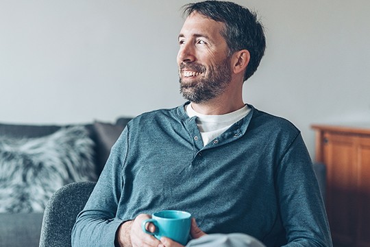 A man with a beard holds a blue mug while smiling, seated in a cozy living room with neutral tones and a plush throw pillow in the background.
