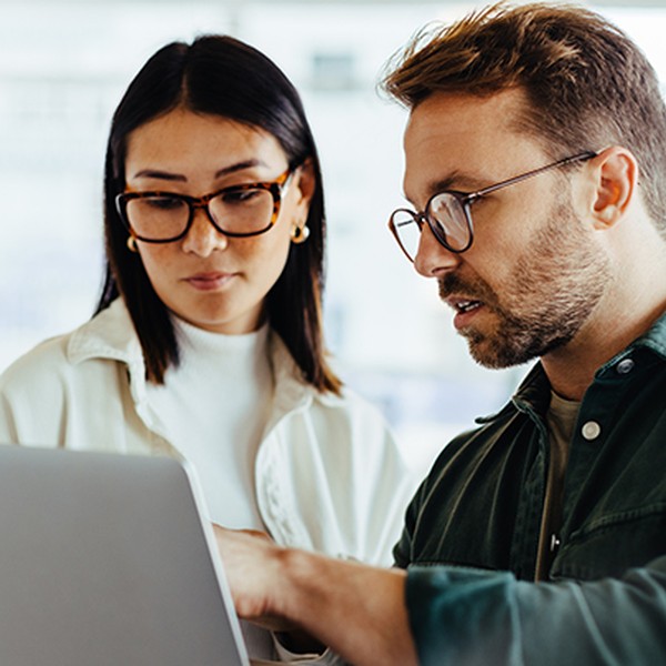 A man and a woman are closely examining a laptop screen together. They appear engaged in discussion, with a bright, modern workspace as their backdrop.