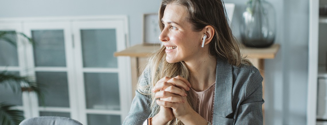A woman with long hair sits at a table, smiling while wearing wireless earbuds. The background features indoor plants and a well-lit, modern workspace with wooden furniture.