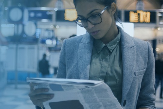 A woman in a gray blazer is reading a newspaper while standing in a busy airport terminal, surrounded by blurred figures and digital displays in the background.