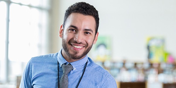 A smiling young man wearing a blue shirt and tie stands confidently in a bright, modern classroom, with bookshelves and colorful artwork visible in the background.