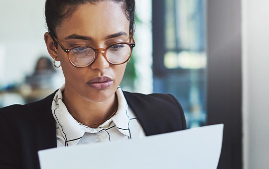 A woman wearing glasses reads a document attentively in a modern office environment. The background features blurred office furnishings, suggesting a professional workspace.