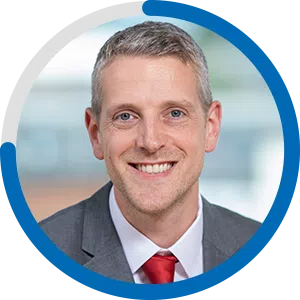 A smiling man in a suit with a red tie is the focus, centered within a circular frame. The background is blurred, suggesting a professional environment.