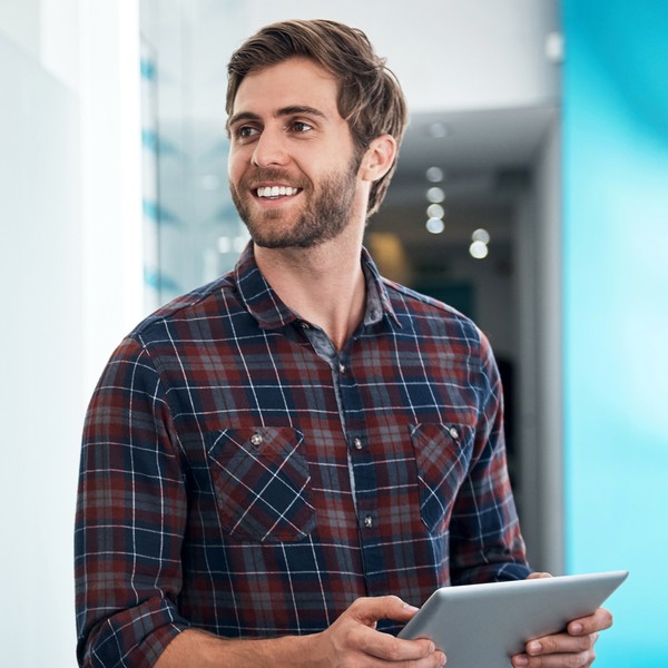 A man wearing a plaid shirt stands holding a tablet, smiling while looking to the side. He is in a brightly lit, modern interior with blue accents and glass features.