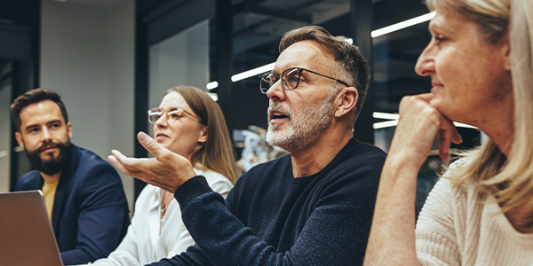 A middle-aged man in glasses gestures while speaking during a meeting. Three colleagues listen attentively, seated at a conference table in a modern office with glass walls.