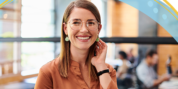 A young woman with glasses and earrings smiles while touching her hair. She stands in a bright, modern café, with people working and socializing in the background.