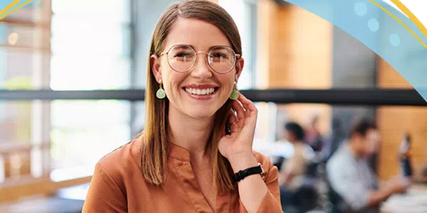 A young woman with glasses and earrings smiles while touching her hair. She stands in a bright, modern café, with people working and socializing in the background.