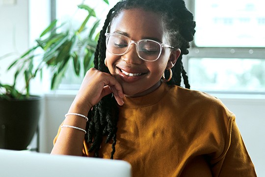 A woman with braided hair and glasses smiles while resting her chin on her hand, looking at a laptop, in a bright room with plants in the background.