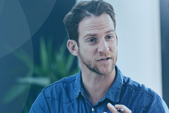 A man in a denim shirt speaks animatedly, gesturing with his hands. A woman listens attentively. They are in a modern office setting with soft lighting and greenery in the background.