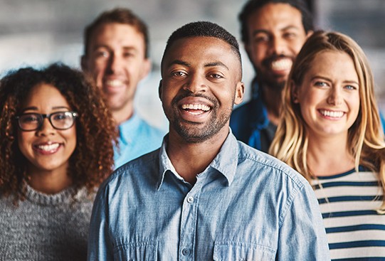 A group of five smiling individuals stands closely together, showcasing diverse hairstyles and clothing styles, against a blurred interior background, conveying a sense of camaraderie and positivity.