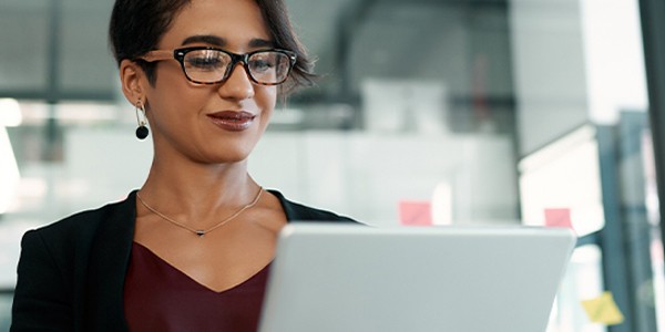 A woman with short hair and glasses is engaging with a laptop. She appears focused and is in a modern office environment with blurred windows and colorful sticky notes in the background.