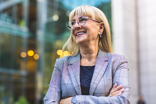 A woman in a plaid blazer stands with her arms crossed, smiling confidently. She is outdoors in a modern urban environment with blurred lights and glass buildings in the background.
