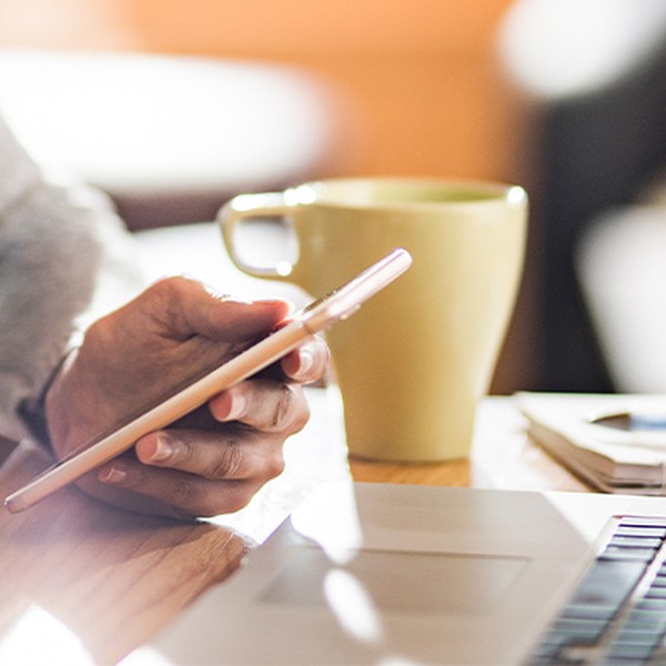 A hand holds a smartphone while resting on a wooden table, next to a green mug and a laptop, suggesting a cozy workspace or café environment.