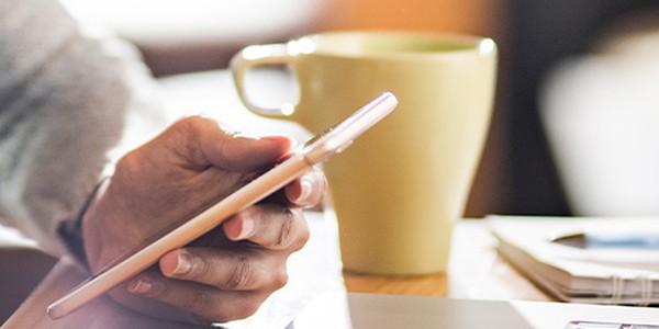 A hand holds a smartphone while resting on a wooden table, next to a green mug and a laptop, suggesting a cozy workspace or café environment.