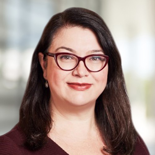 A woman with long, dark hair and glasses smiles gently. She appears in a professional setting, with a softly blurred background suggesting an office environment.