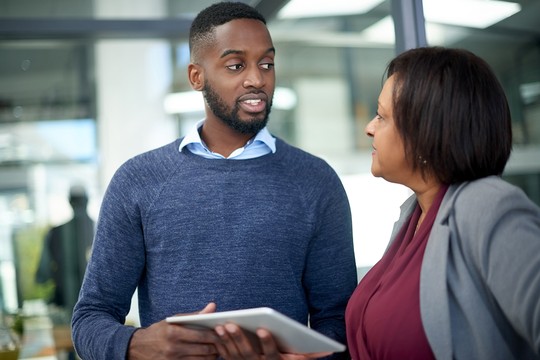 A man holds a tablet while engaged in conversation with a woman in a professional setting. They appear to be discussing something important, with modern office elements in the background.
