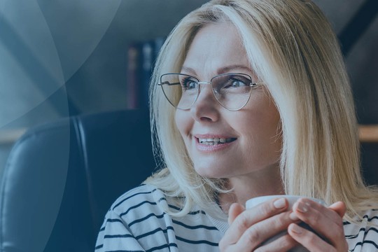 A woman with long, blonde hair and glasses smiles while holding a coffee cup. She sits in a comfortable chair, surrounded by a cozy, modern room with plants and shelves.