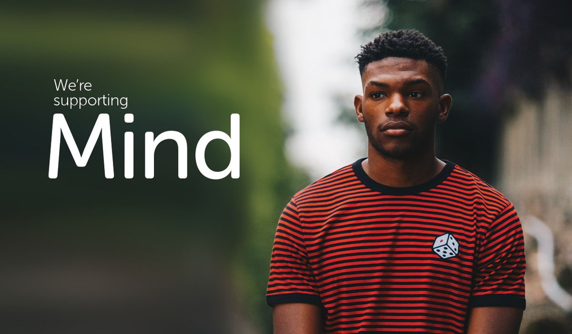 A young man wearing a red and black striped shirt stands confidently, gazing forward. Behind him is a blurred outdoor setting, suggesting a casual urban environment. 

Text: "We're supporting Mind"