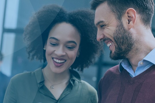 Two professionals, a woman and a man, are smiling and looking at a device together, engaged in conversation. The background features other colleagues in an office setting.