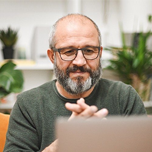 A middle-aged man with a beard and glasses smiles while clasping his hands, focused on a laptop. He sits on a couch surrounded by indoor plants in a bright, cozy space.