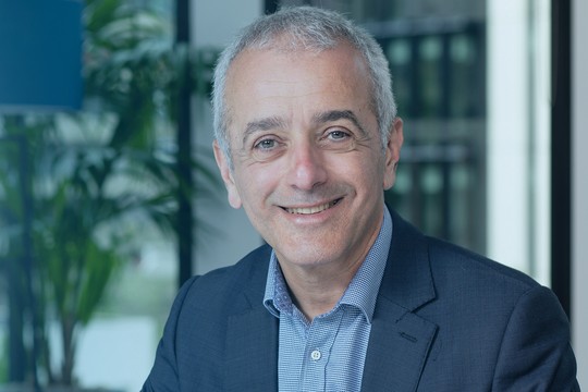 A man in a suit smiles while sitting in an office with large windows. Background features greenery and soft blue tones. The atmosphere is professional and welcoming.