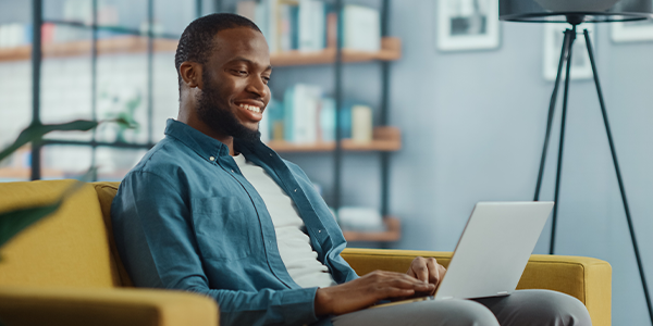 A man seated on a yellow couch engages with a laptop, smiling. Behind him are bookshelves and a lamp, suggesting a cozy, modern living space.