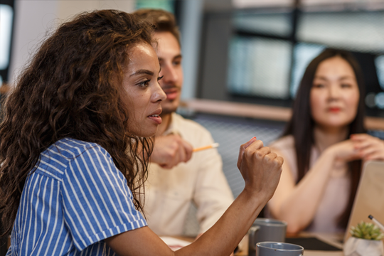 A woman with curly hair, wearing a striped shirt, gestures expressively while engaged in discussion with two seated colleagues in a modern workspace, surrounded by cups and a laptop.