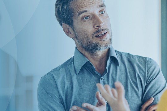 A man in a blue shirt gestures with his hands while speaking to a person out of view. The setting is bright and modern, suggesting a professional environment.