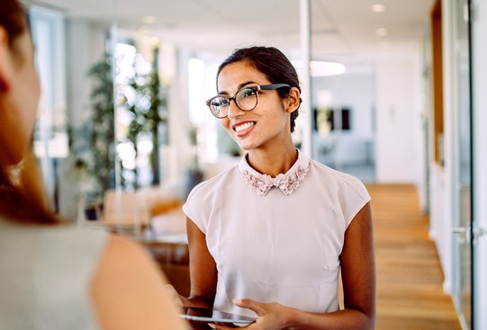 A young woman in a blouse and glasses smiles while holding a tablet, engaging in conversation with another person, set in a modern office with large windows and greenery.