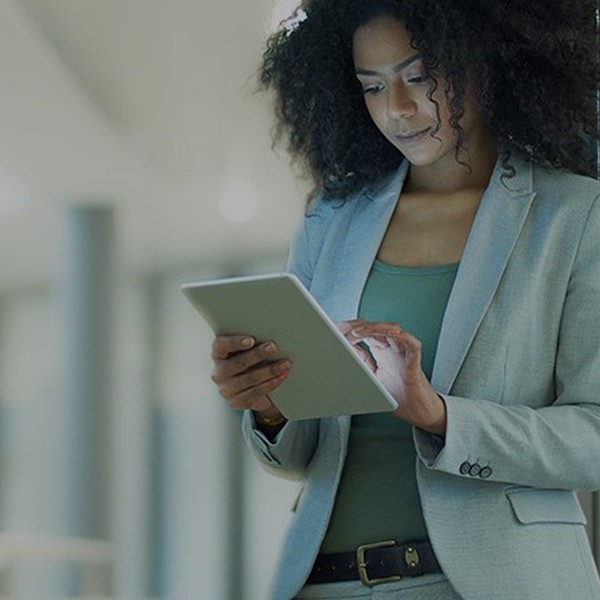 A woman in a light gray suit is intently using a tablet while leaning against a wall in a modern, well-lit environment with blurred backgrounds suggesting an office setting.