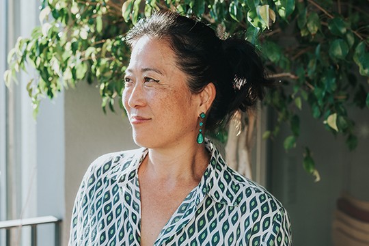 A woman with dark hair tied back wears a patterned shirt and green earrings. She gazes thoughtfully to the side, surrounded by lush greenery in a bright, well-lit environment.