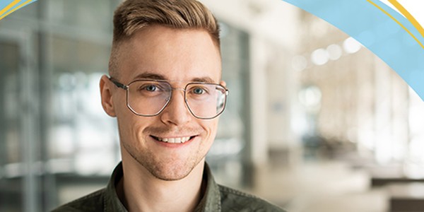 A young man with glasses and styled hair smiles while standing in a modern indoor space with blurred background features, such as glass walls and seating areas.