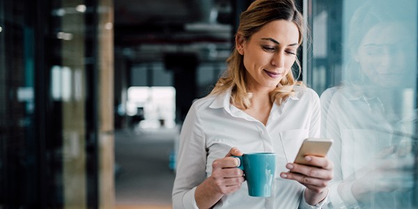 A woman holds a teal mug and looks at her smartphone with a smile. She stands by a glass wall in a modern office environment, with blurred office elements in the background.