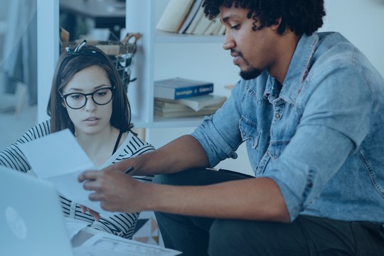 A laptop sits on a table as two people review papers together. They are engaged in conversation, surrounded by a cozy indoor space with plants and books in the background.