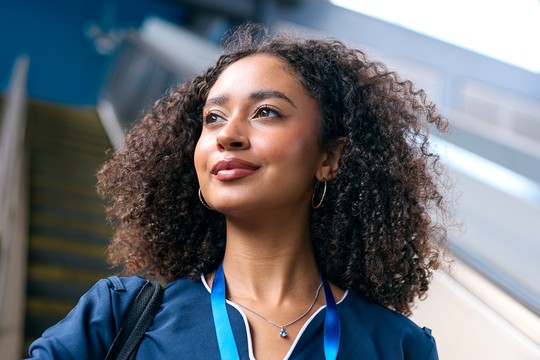 A young woman with curly hair gazes upward, wearing a blue outfit and a necklace, with a lanyard around her neck. Stairway and bright surroundings hint at a public space.