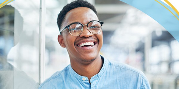 A young man with glasses smiles broadly, leaning against a glass surface in a bright, open environment filled with soft lighting and blurred background details.