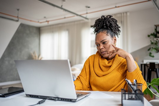 A woman in a bright orange sweater thoughtfully engages with a laptop on a desk surrounded by a well-lit, modern home office. Papers and office supplies are nearby.