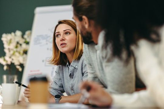 A woman is speaking intently in a meeting, holding a pen, while two people listen attentively. A coffee cup and flowers are visible on the table in a modern setting.