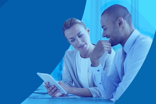 Two professionals, a woman and a man, are discussing while examining a tablet in a modern office setting. The background features various shades of blue, creating a calm atmosphere.