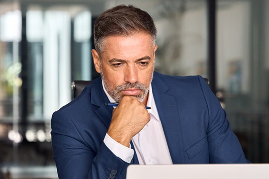A man in a blue suit sits thoughtfully at a desk, resting his chin on his hand, focused on a laptop in a modern office with blurred glass windows in the background.