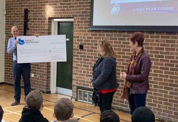 A man holds a large check promoting a school donation in front of two women. They stand in a school gym with a brick wall and projector screen behind them, while students observe.