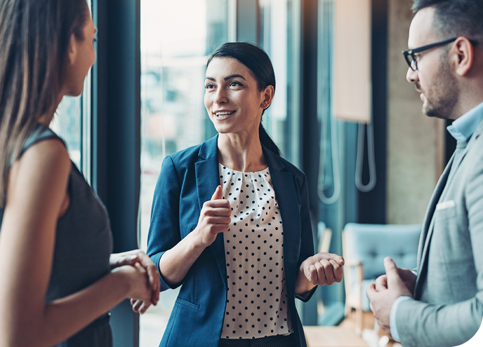 A woman in a blazer is engaging in conversation with two individuals, smiling and gesturing with her hands. They stand in a modern office with large windows and contemporary furnishings.