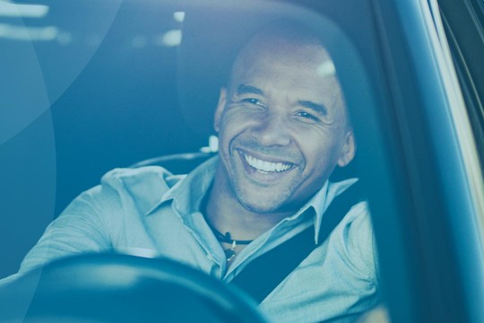 A man smiles while seated in a car, resting his arm on the open window. The environment appears bright and cheerful, suggesting a pleasant day.