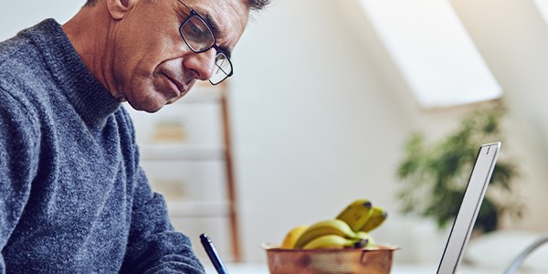 A man wearing glasses writes in a notebook while focused on a laptop, surrounded by a bright, airy room with a fruit bowl containing bananas in the foreground.