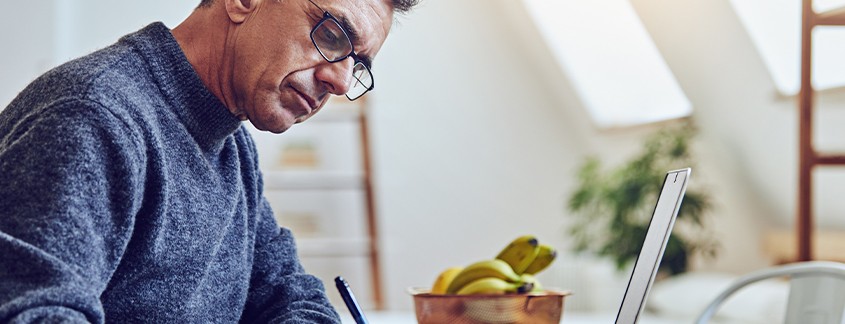A man wearing glasses writes in a notebook while focused on a laptop, surrounded by a bright, airy room with a fruit bowl containing bananas in the foreground.