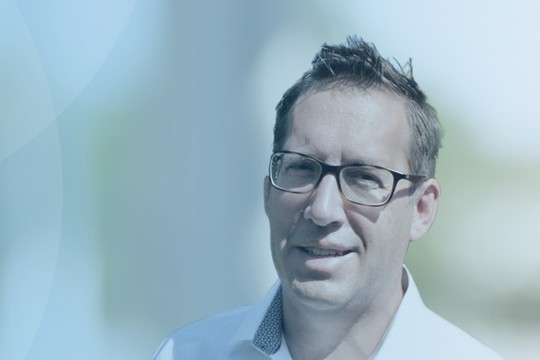 A person wearing glasses smiles, standing against a softly blurred light blue background, suggesting an outdoor setting. The scene conveys a calm and professional atmosphere.