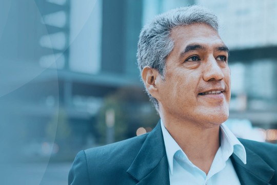 A man in a suit stands with a slight smile, looking upward, in a modern urban environment with blurred buildings and greenery in the background. The atmosphere is optimistic and professional.