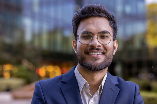A man with glasses and a beard smiles confidently while wearing a blazer. He stands outdoors in front of a building with large windows, surrounded by greenery and soft, blurred lights.